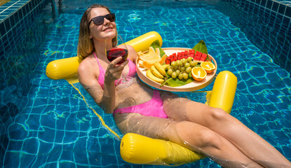 Beautiful woman lying on floating hammock in the swimming pool with wine and tray of fruits - 790608752