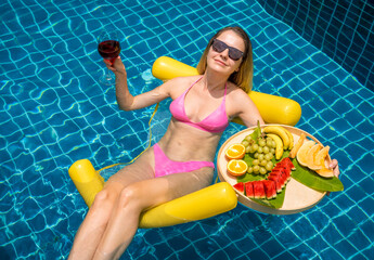 Beautiful woman lying on floating hammock in the swimming pool with wine and tray of fruits - 790612175