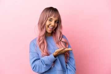 Young mixed race woman with pink hair isolated on pink background presenting an idea while looking...