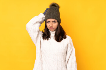 Young woman with winter hat over isolated yellow background having doubts while scratching head
