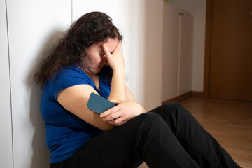 Single sad woman holding a mobile phone lamenting sitting on the floor in her home with a dark light in the background