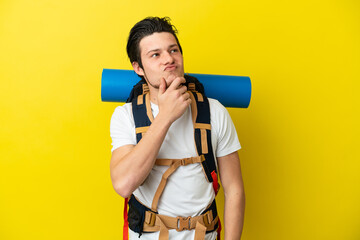Young mountaineer Russian man with a big backpack isolated on yellow background having doubts