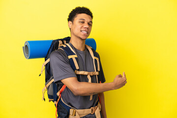 Young mountaineer African American man with a big backpack isolated on yellow background pointing...