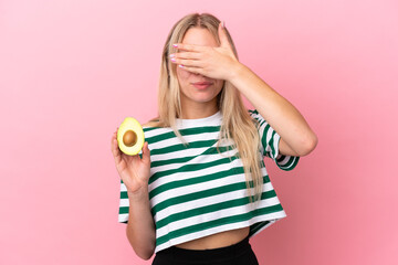 Young caucasian woman holding an avocado isolated on pink background covering eyes by hands. Do not...
