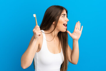 Young caucasian woman brushing teeth isolated on blue background shouting with mouth wide open to...