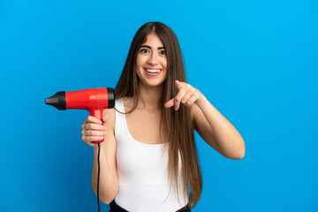 Young caucasian woman holding a hairdryer isolated on blue background surprised and pointing front