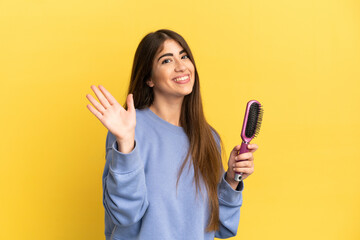Young caucasian woman holding hairbrush isolated on blue background saluting with hand with happy...