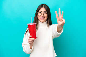 Young caucasian woman drinking soda isolated on blue background happy and counting three with...