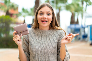 Young pretty blonde woman holding a wallet at outdoors surprised and pointing finger to the side