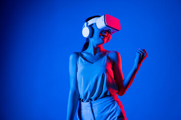 Smart female standing wearing VR headset connecting metaverse, future cyberspace community technology. Woman victory competition. Lady gesticulate raising arm slightly with happy face. Hallucination. - obrazy, fototapety, plakaty