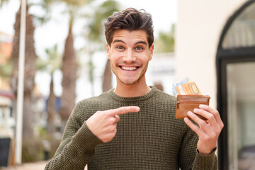 Young caucasian man at outdoors holding wallet with money and pointing it