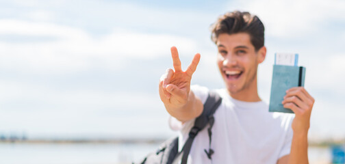Young caucasian man holding a passport at outdoors smiling and showing victory sign