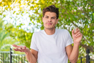 Young caucasian man holding invisible braces at outdoors making doubts gesture while lifting the shoulders