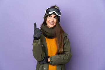 Skier caucasian girl with snowboarding glasses isolated on purple background pointing up a great...