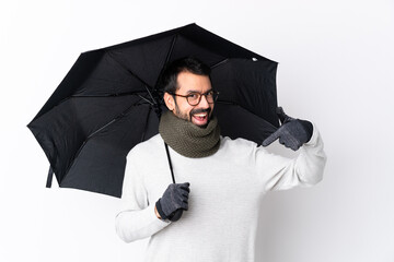 Caucasian handsome man with beard holding an umbrella over isolated white wall proud and...