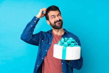 Young handsome man with a big cake over isolated blue background having doubts while scratching head