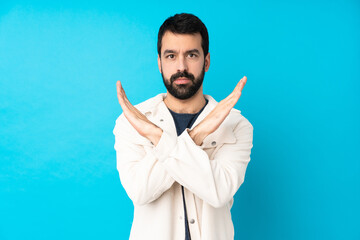 Young handsome man with white corduroy jacket over isolated blue background making NO gesture