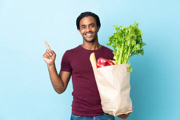 African American man holding a grocery shopping bag isolated on blue background showing and lifting...