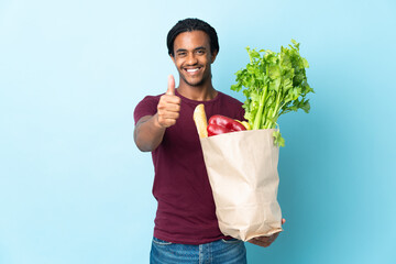 African American man holding a grocery shopping bag isolated on blue background with thumbs up...