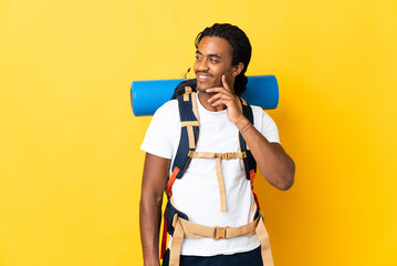Young mountaineer man with braids with a big backpack isolated on yellow background thinking an...