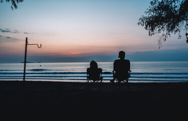 A man and a teddy bear sit on a chair and watch the sunrise on the beach.