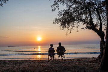 Tourist couple on the beach under the tree and watch the sunrise,  colorful golden morning light, Outdoor lifestyle concept.