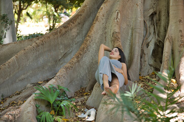South American woman, young, pretty, brunette with gray top and skirt lying on the roots of a big tree, relaxed and calm. Concept beauty, fashion, diversity, peace, nature.