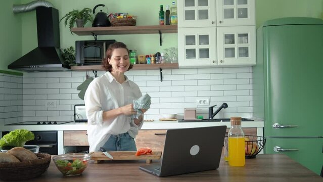 A young Caucasian woman is having fun and dancing while cooking in a brightly lit kitchen.