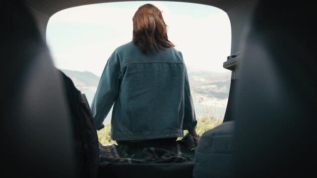 An adult woman enjoying a beautiful sea view while sitting in the trunk of her SUV car.