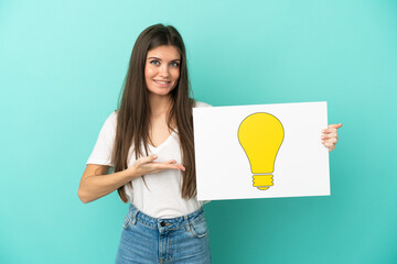Young caucasian woman isolated on blue background holding a placard with bulb icon and pointing it - 790669159