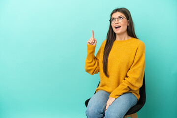 Young caucasian woman sitting on a chair isolated on blue background pointing up and surprised - 790669167