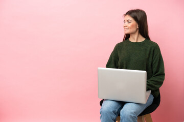 Young caucasian woman sitting on a chair with her laptop isolated on pink background looking side - 790669185