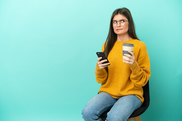 Young caucasian woman sitting on a chair isolated on blue background holding coffee to take away and a mobile while thinking something - 790669189