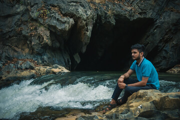 Young man sitting on the edge of a cave through which water stream ias flowing, enjoying the view.