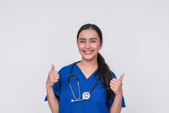 Young Asian female caregiver in blue scrub giving thumbs up, isolated on white