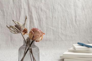 Gray fabric space with a vase of dried rose flowers, a blue pen and a stack of white notebooks	