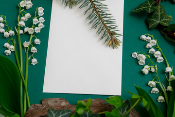Composition on a table with white blank sheet of paper, surrounded by ivy plants and lilies of the valley.