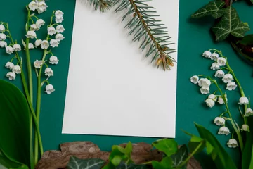 Poster Composition on a table with white blank sheet of paper, surrounded by ivy plants and lilies of the valley. © Katarzyna