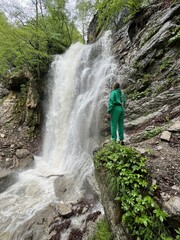 Obraz premium A woman on the background of the Lyazhginsky waterfall in the spring forest. A cascading waterfall surrounded by greenery. Splashes of water. The water flows along the Lyazhgi River. Ingushetia