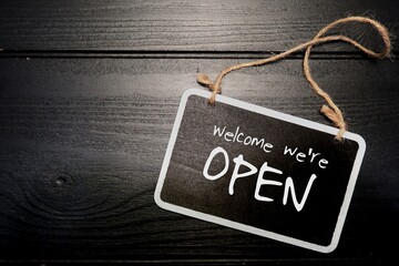 Small black chalkboard sign on black wood background with text handwritten Welcome We're Open,...