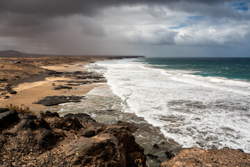 A rocky beach with a cloudy sky and a large body of water. The sky is overcast and the waves are...