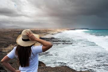 A woman wearing a straw hat stands on a rocky beach looking out at the ocean. The sky is cloudy and...