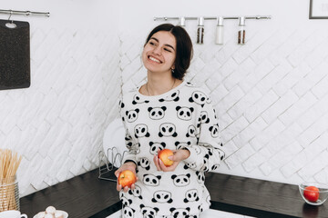 Cheerful woman standing in bright kitchen smiling laughing eating apple fruit, dressed in patterned pajama set. The kitchen counter arranged with ingredients, utensils, atmosphere domestic