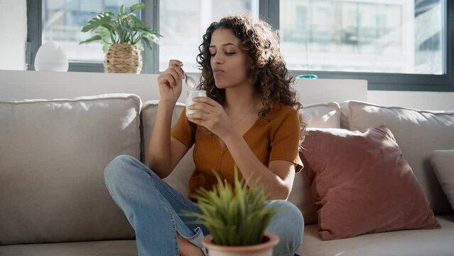 Video of beautiful woman eating healthy yogurt while sitting on couch at home.