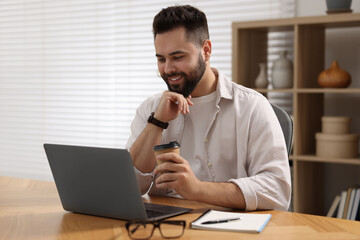 Young man with cup of coffee watching webinar at table in room