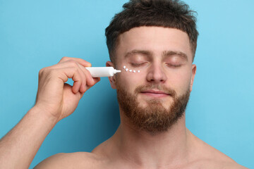 Handsome man applying moisturizing cream onto his face on light blue background