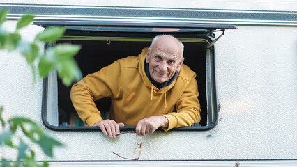 Cheerful senior man peering out from camper window holding glasses in hand. Joyful retirement road trip vibes