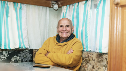 Smiling elderly man with crossed arms sitting in cozy camper interior. Comfortable retirement travel lifestyle