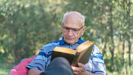 Engrossed elderly man reading book in peaceful outdoor setting surrounded by trees. Intellectual leisure in nature embrace enhances wellbeing and serenity