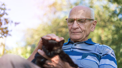 Content senior man with glasses holds small black cat, bonding moments in sunny garden. Gentle companionship and outdoor relaxation captured in serene setting